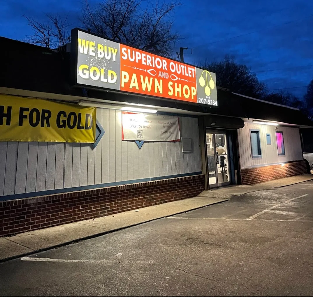 Superior Outlet and Pawn Shop at dusk on Richmond Highway: a lit sign reading We Buy Gold above the entrance, a yellow Cash For Gold banner along the side wall, and the parking lot in front.